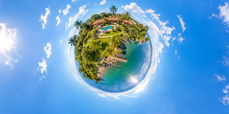 A sunny tiny-planet panorama of a lakeside resort with tropical greenery, warm roofs and bright blue sky.